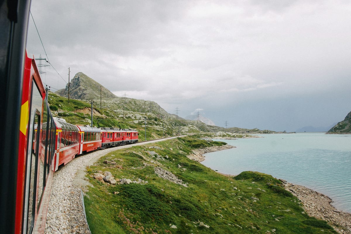 A Bernina Express train with panoramic carriages runs beside a pale blue mountain lake under grey clouds, seen from the train window.