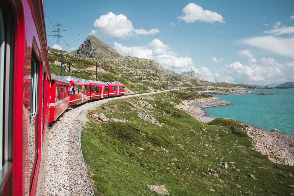 A red train curves along the turquoise shore of a mountain lake under a summer sky, seen from the train window.