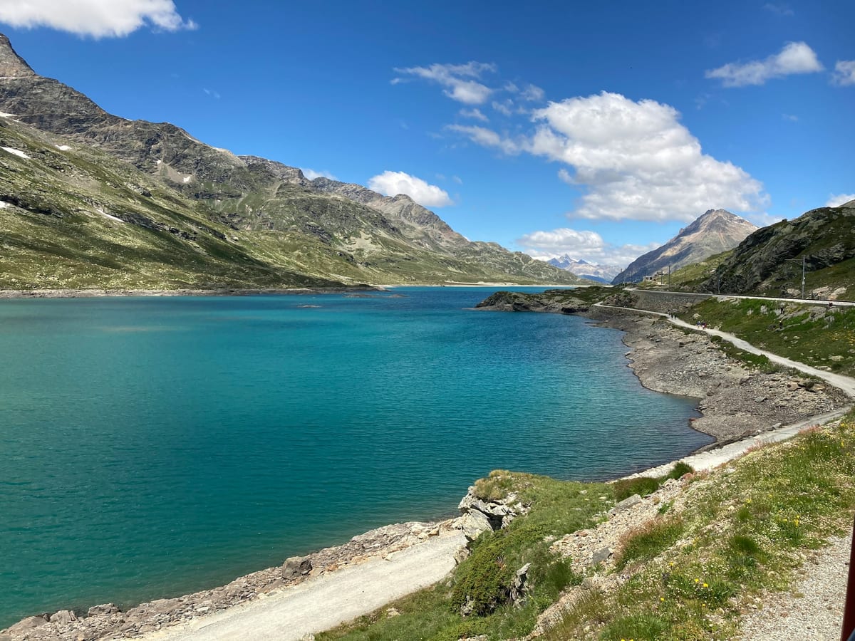 Turquoise Lago Bianco at the Bernina Pass under a blue summer sky, with green mountain slopes on both sides.