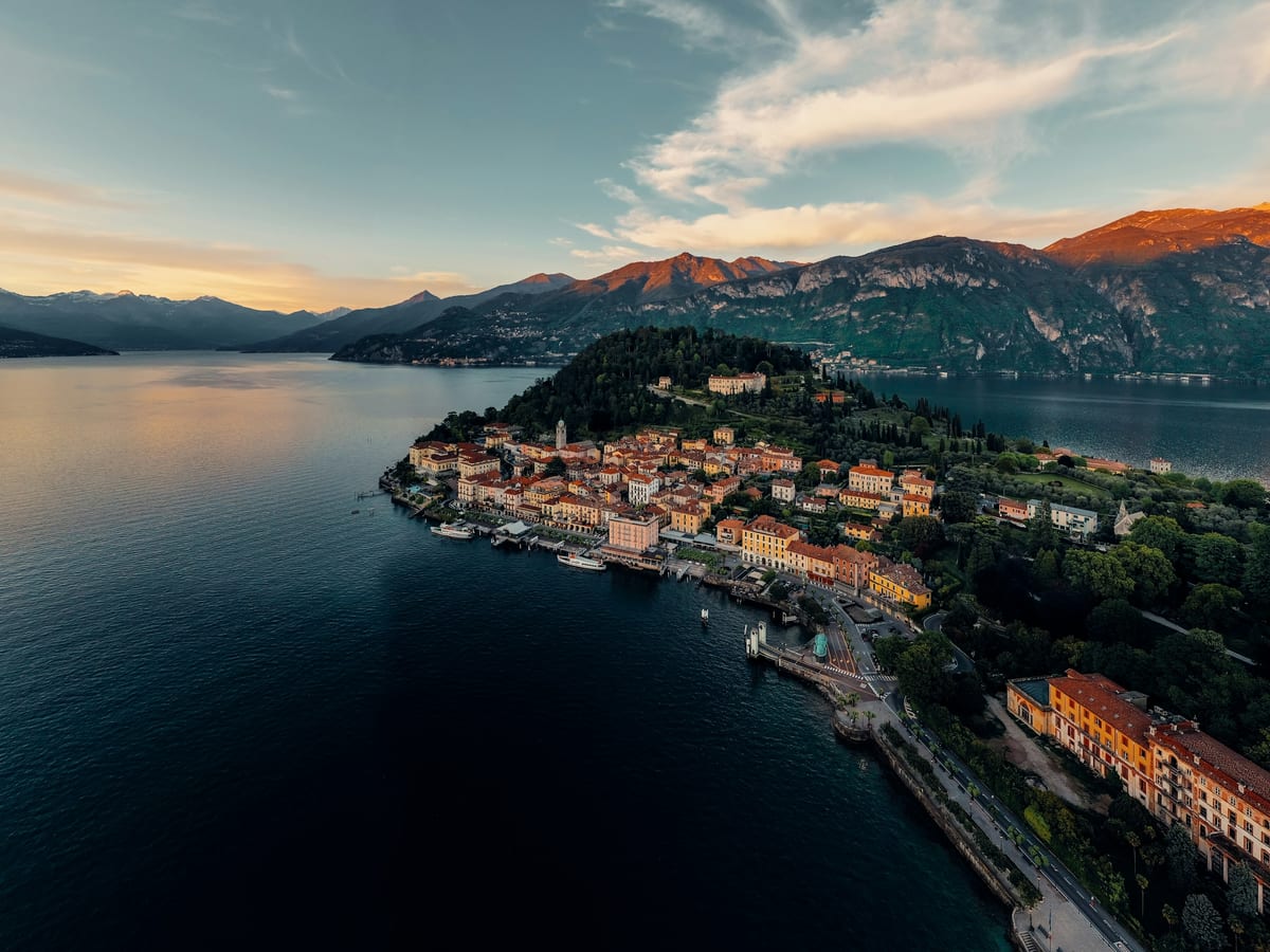 Aerial view of a lakeside town on a wooded point of Lake Como in the warm light of a low sun, with the lake on both sides and mountains beyond.