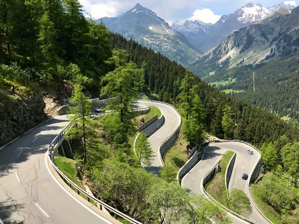 The Maloja Pass road zigzags down through larch forest in tight hairpin bends, with rocky peaks beyond.