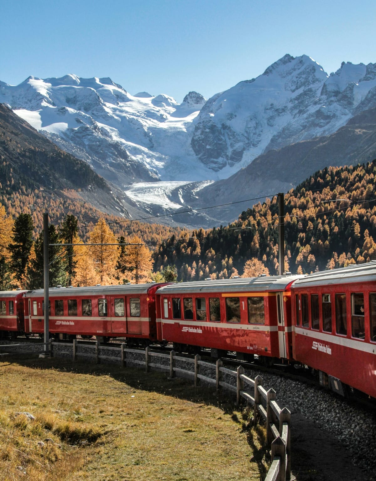 A red train curving past autumn larches beneath snowy peaks and the Morteratsch Glacier.