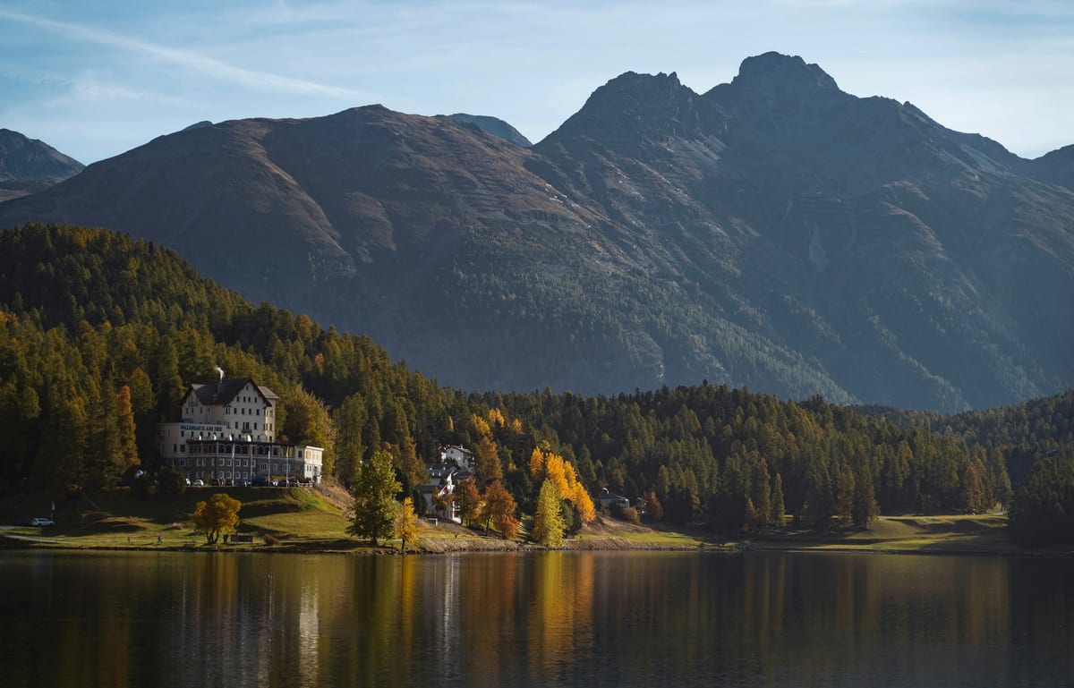 Autumn larches and a lakeside building reflected in calm water at St. Moritz, below a dark mountain ridge.